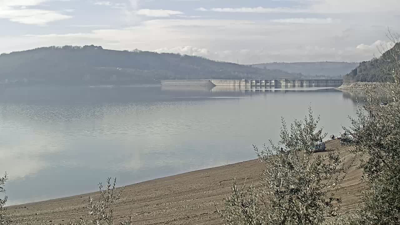 A calm lake with a dam and low, tree-covered hills in the background under a partly cloudy sky.