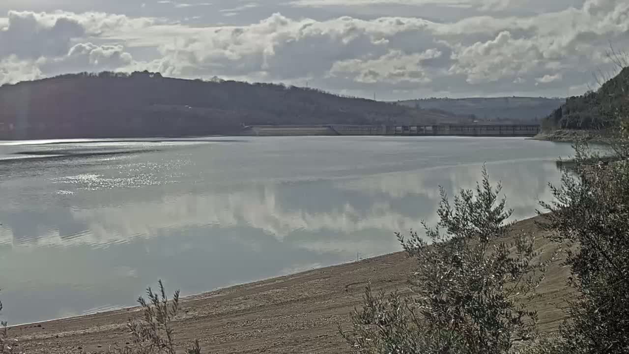 A calm body of water reflects a cloudy sky, with a dam visible in the distance and a sandy shoreline in the foreground.