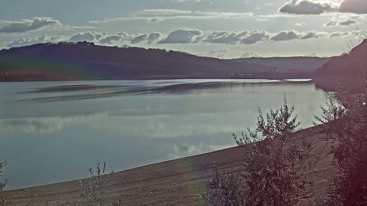 A calm lake reflects a cloudy sky under mostly sunny conditions, with hills in the background and sparse vegetation in the foreground.