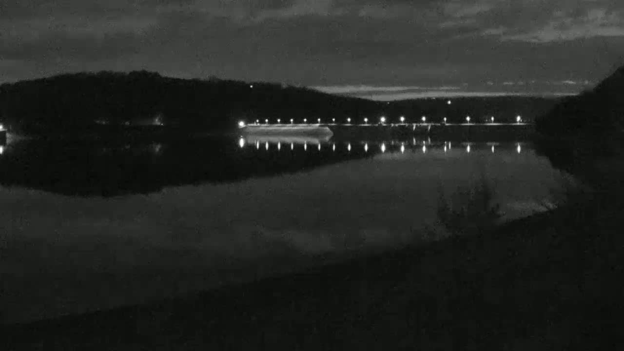 A calm, dark lake reflects the lights of a distant dam under a nighttime sky.