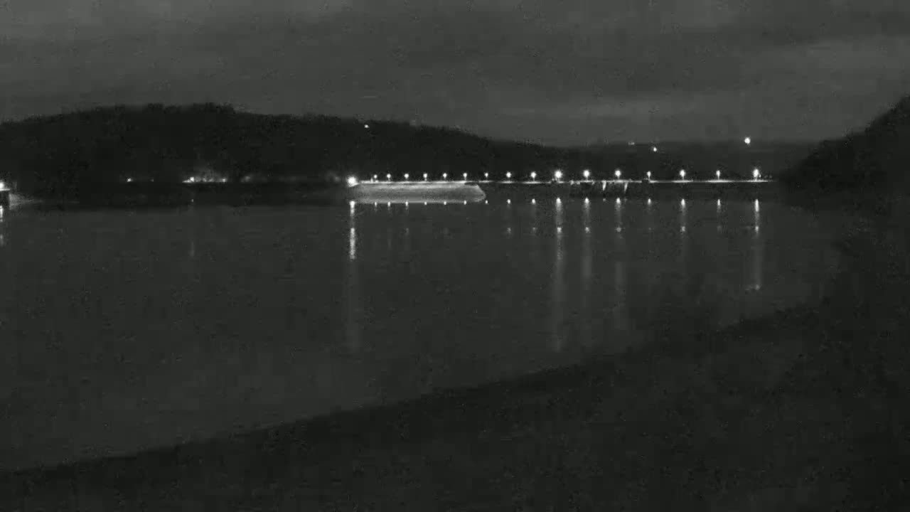 A nighttime view of a calm body of water reflecting the lights of a dam and surrounding hills under a dark sky.