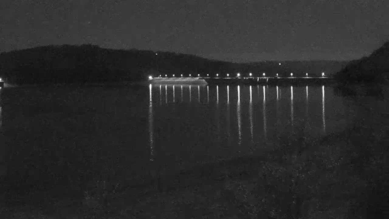 A dark, calm body of water reflects the lights of a dam at night, with hills visible in the background under a clear sky.