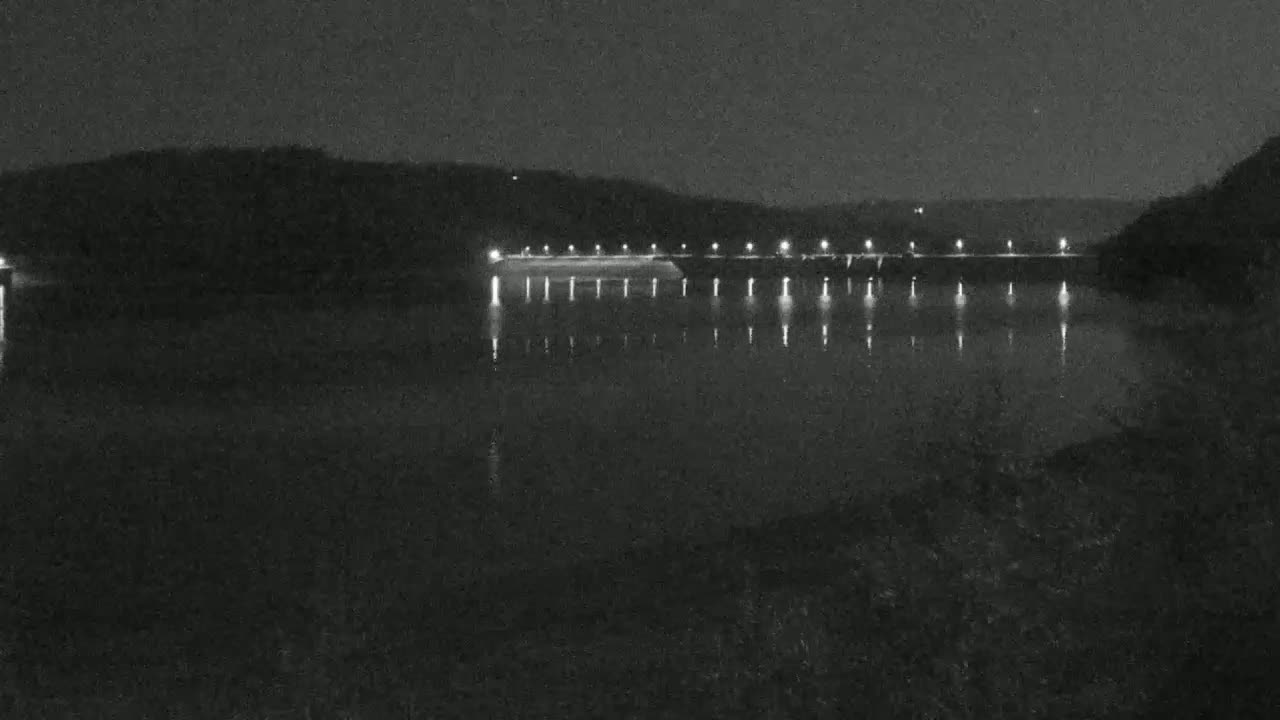 A dark, nighttime scene shows a lit dam across a calm body of water, with hills in the background.