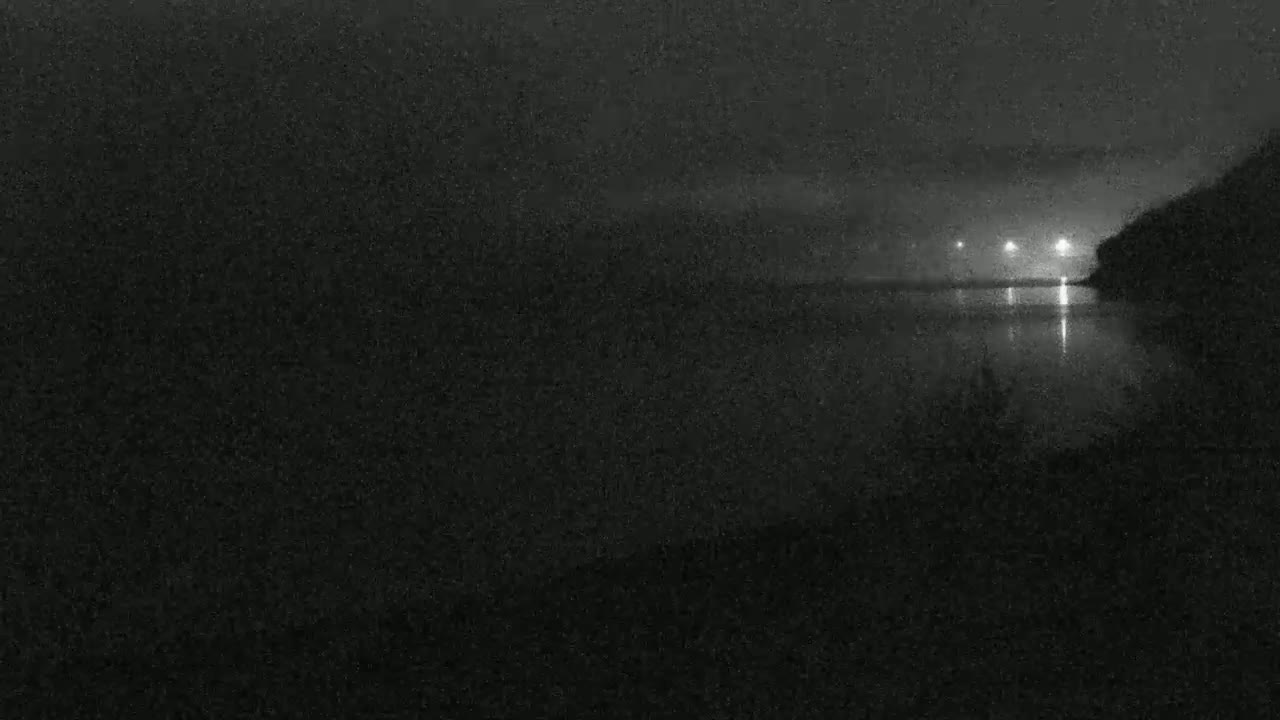 A nighttime view of a dark body of water reflecting distant lights, under a cloudy sky.