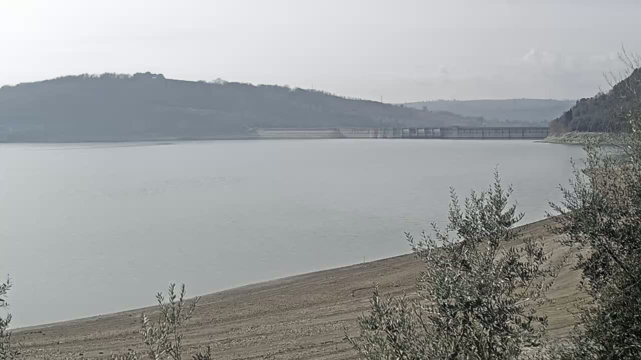 A calm, gray lake with a dam and hills in the background on an overcast day.