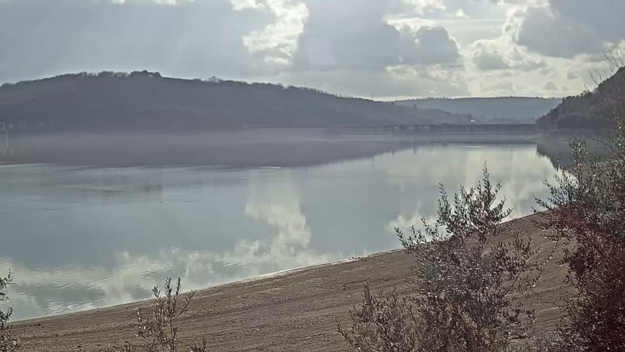 A calm lake reflects a partly cloudy sky, with hills visible in the background and low-lying vegetation in the foreground.