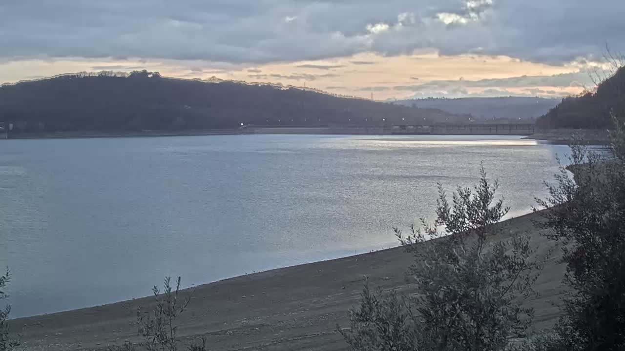 A calm lake under a cloudy sunset sky is bordered by a receding shoreline and hills covered in dark trees.