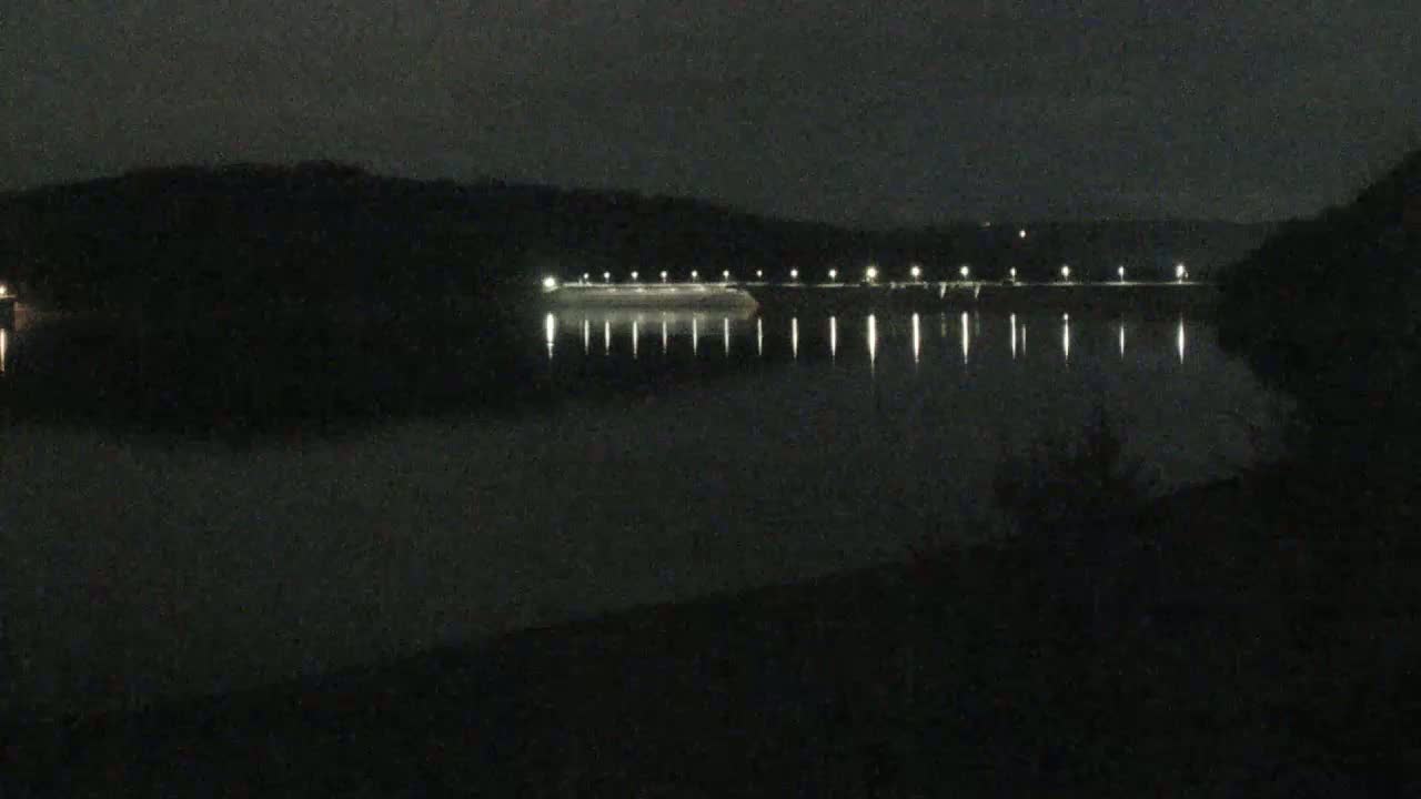 At night, a calm body of water reflects the lights of a dam stretching across its width, under a dark sky.