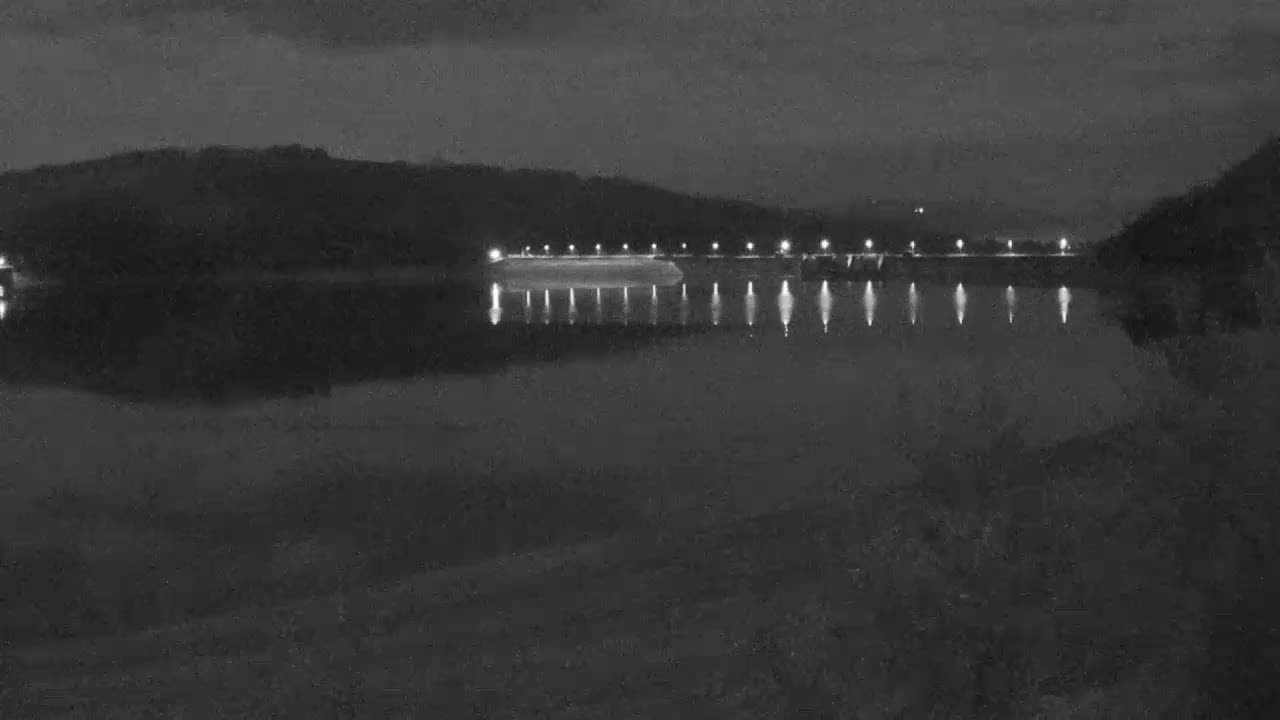 A dark, nighttime scene shows a body of water reflecting the lights of a structure along the far shore, which is backed by a line of dark hills.