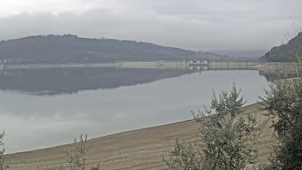 A calm lake reflects a dam and distant hills under an overcast sky.