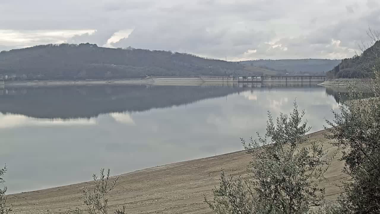 A calm lake reflects a cloudy sky and wooded hills under an overcast sky.