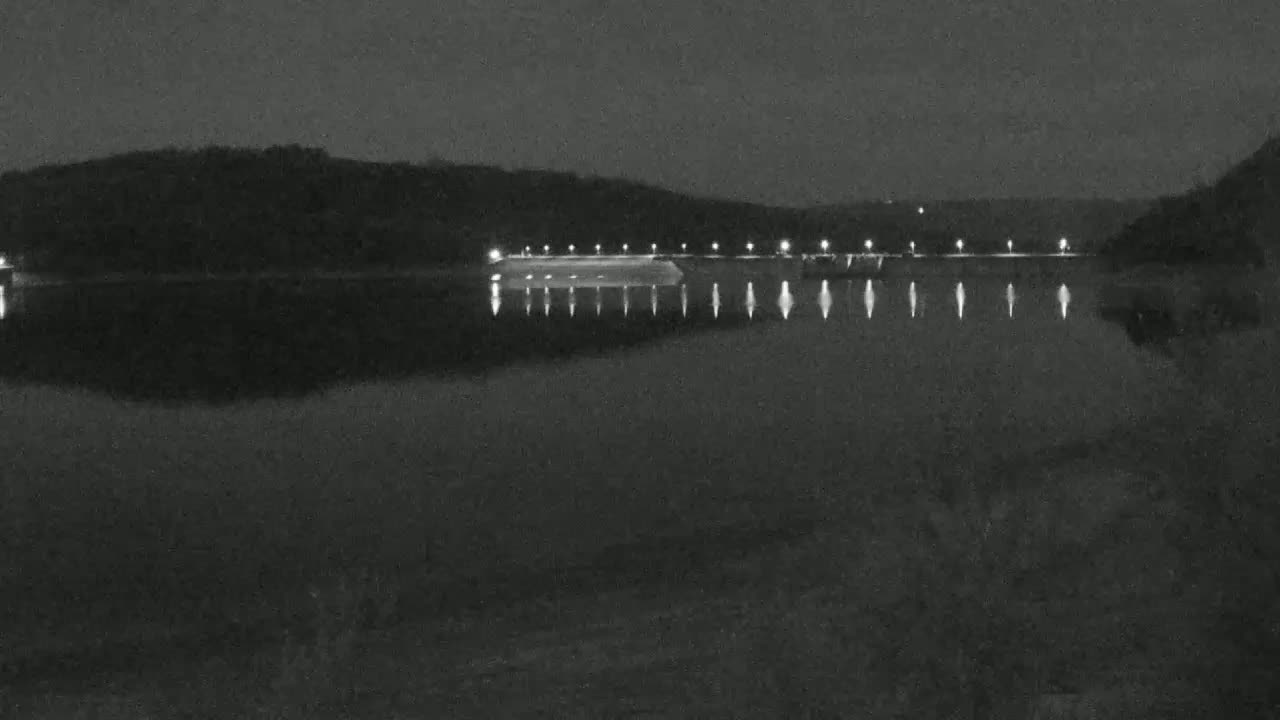 A dark, nighttime view shows a calm body of water reflecting the lights of a structure along the far shore, backed by a treeline.