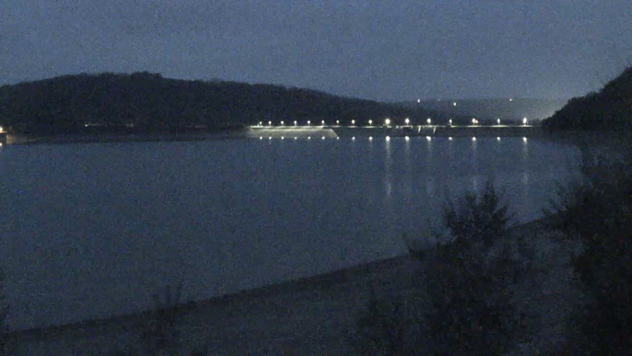 A dark, calm lake reflects the lights illuminating a dam at night, with hills visible in the background under a dusky sky.