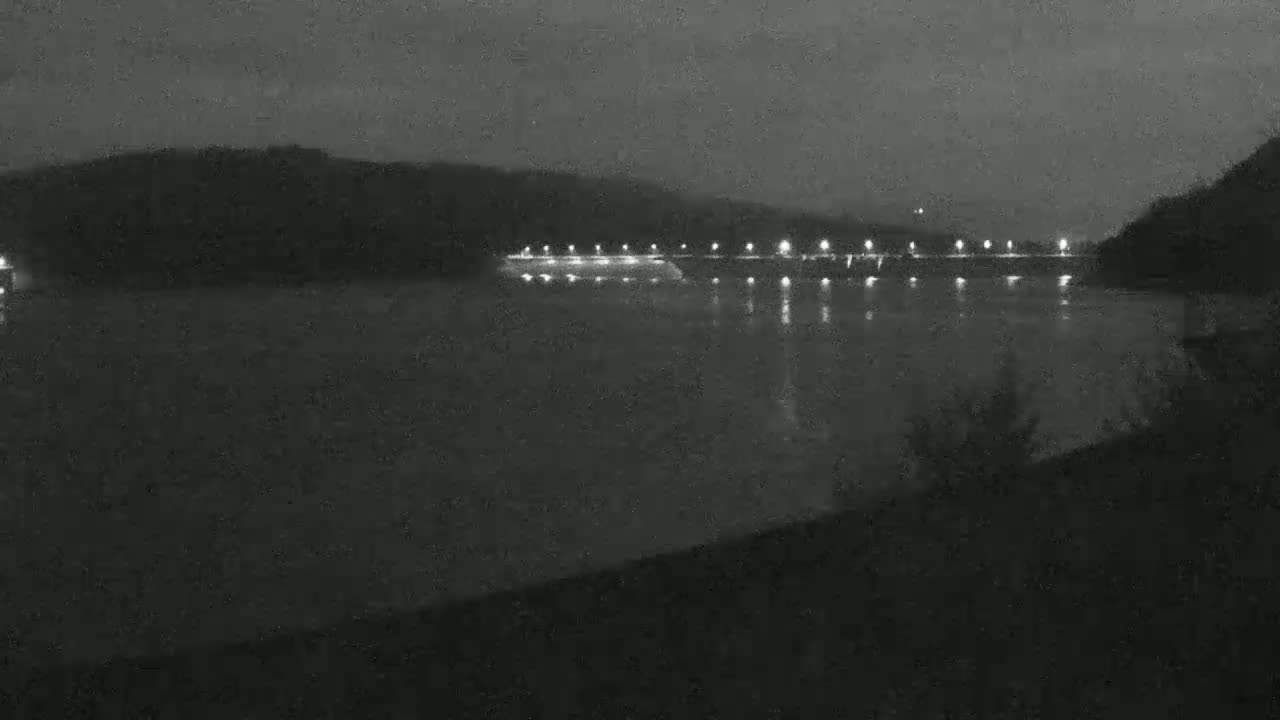 A nighttime view of a dark body of water reflecting the lights of a distant structure on the opposite shore, under a cloudy sky.