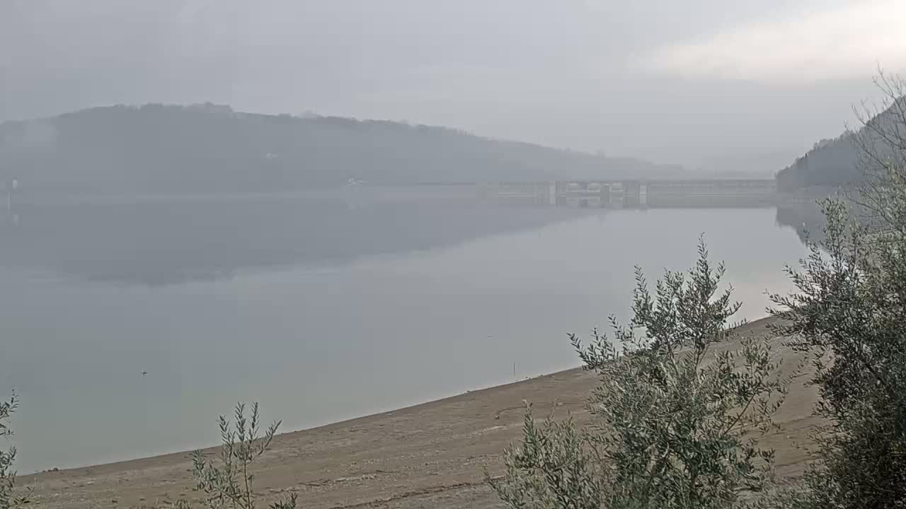 A calm, foggy lake stretches out before a dam and distant hills.