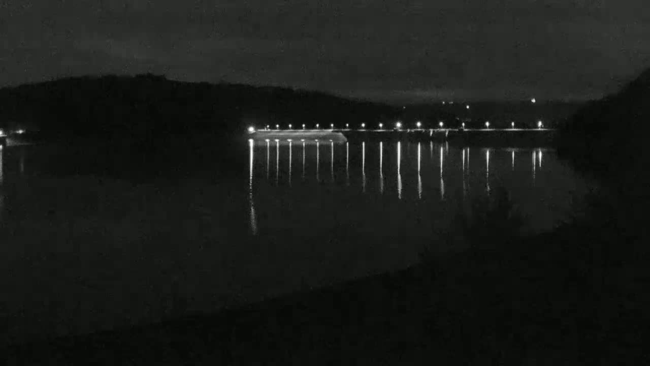 A nighttime view of a calm body of water reflecting the lights of a dam situated against a dark, hilly background.