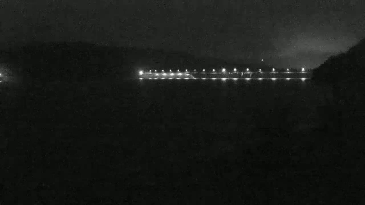 At night, a line of lights illuminates a long structure, possibly a dam or bridge, against a dark, hilly background.