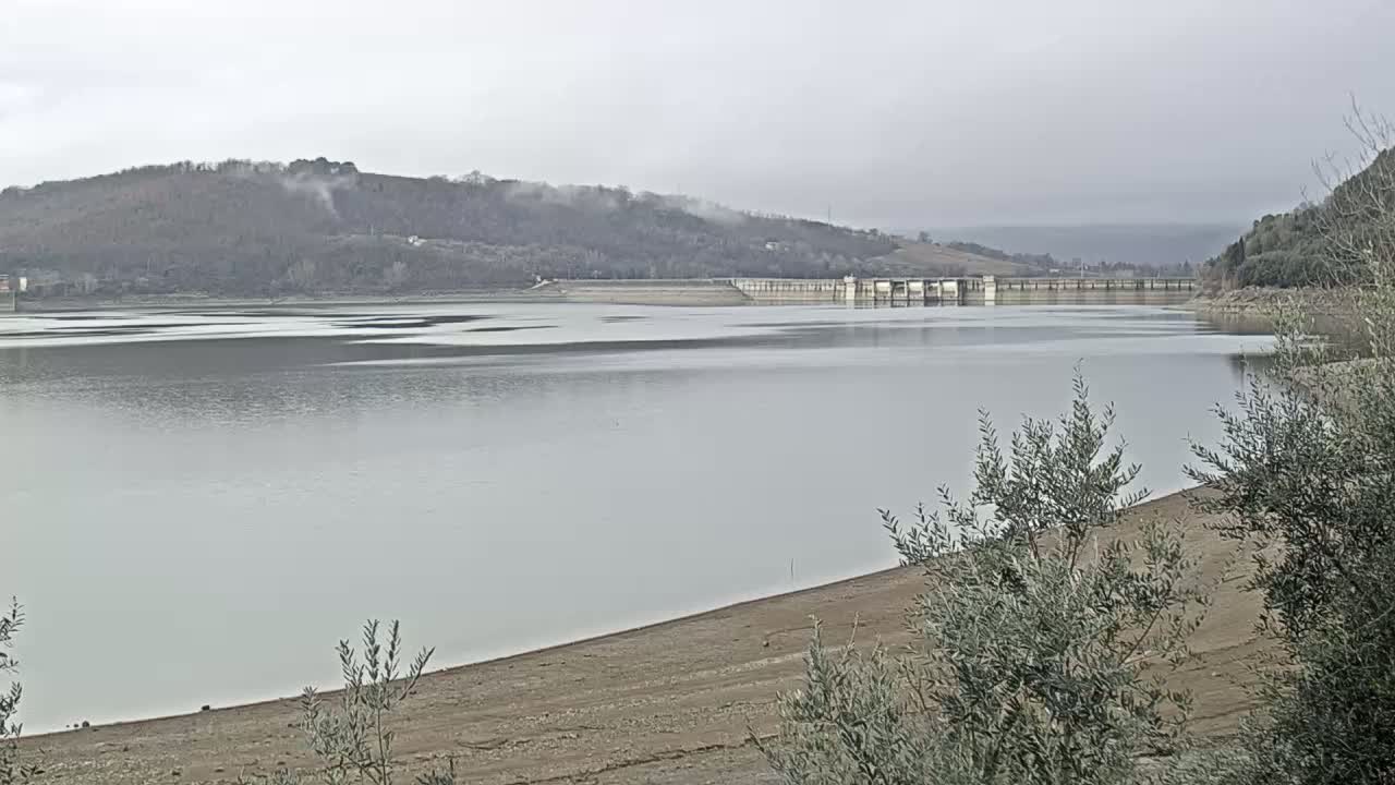 A low-water reservoir with a dam in the background, under an overcast sky.