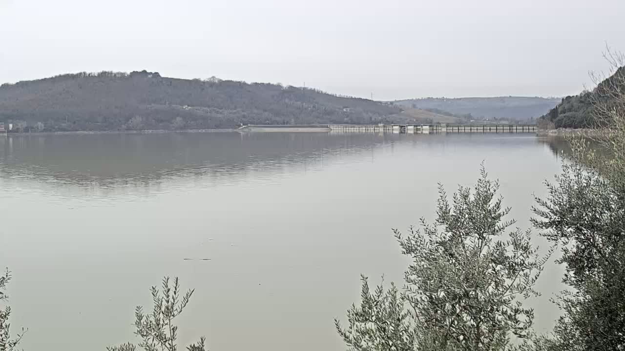 A calm lake stretches out before a hilly landscape under an overcast sky, with a dam and spillway visible in the mid-ground.