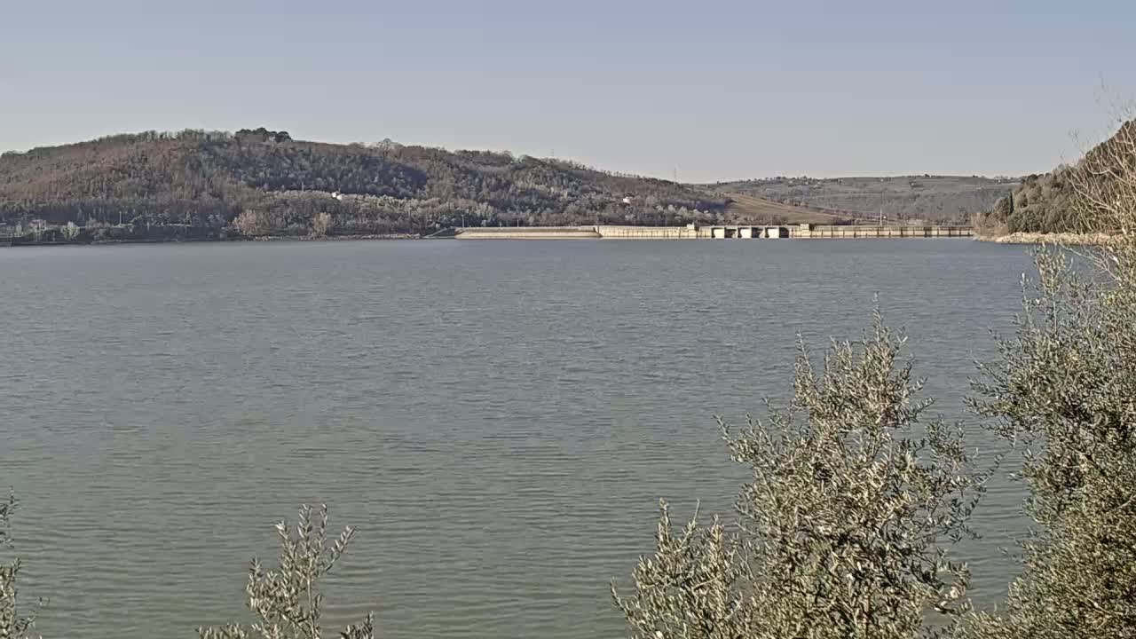 A calm lake stretches out before a dam, with hills covered in sparse, winter trees in the background under a clear sky.