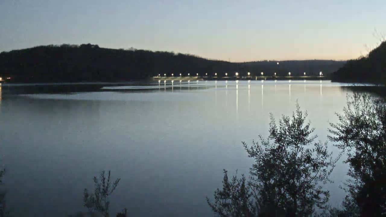 A calm lake at twilight is illuminated by lights across a distant dam, framed by dark trees in the foreground.