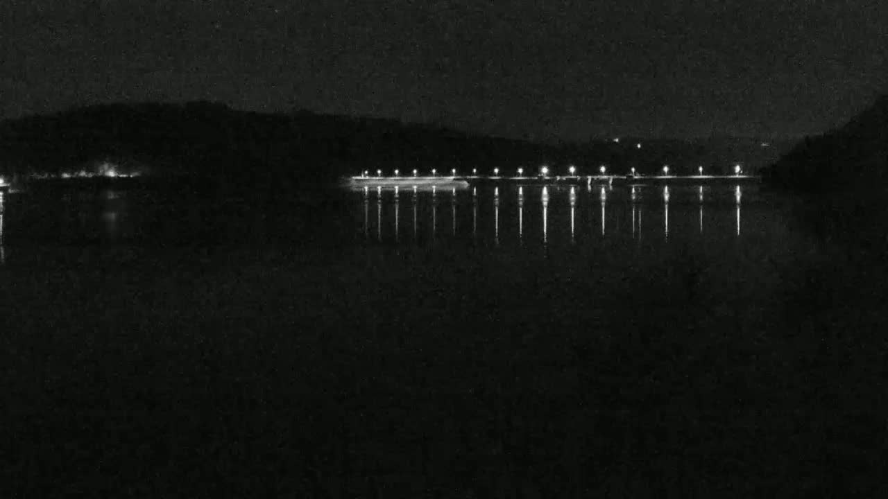 At night, a calm body of water reflects the lights of a long, distant structure on a dark shoreline.