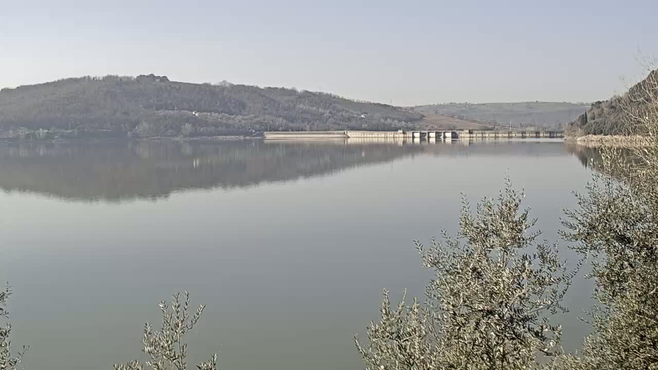 A calm lake reflects a dam and tree-covered hills under a clear sky.