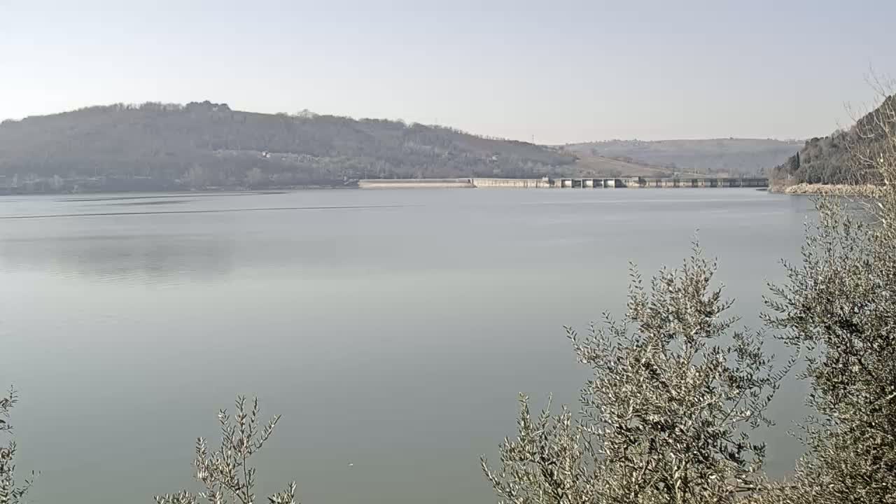 A calm lake stretches out before a dam and hills under a clear, bright sky.