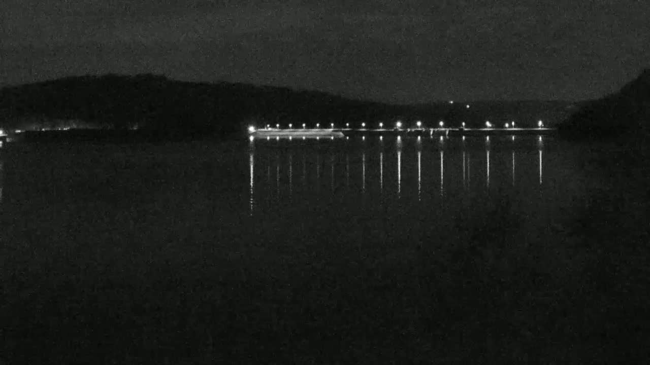 At night, a long structure with lights is reflected in a calm body of water, with dark hills visible in the background.