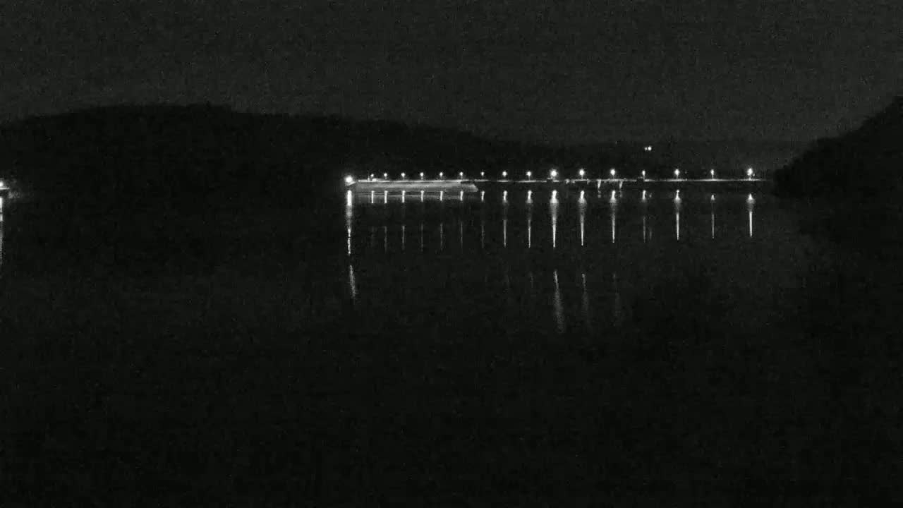 At night, a long, dark structure with lights stretches across a calm body of water, reflecting the lights.