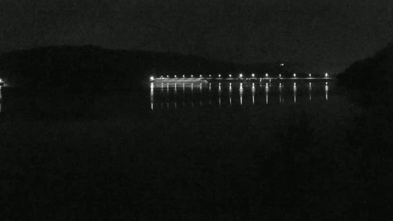 A dark, calm body of water reflects the lights of a long, low structure on the far shore at night.