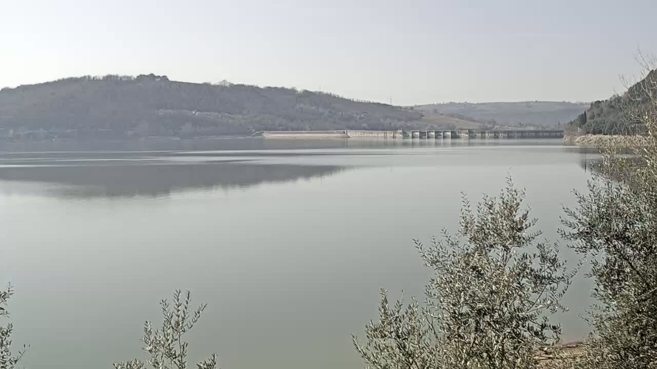 A calm lake reflecting a hazy sky stretches before a dam and hillsides covered in sparse, winter trees.