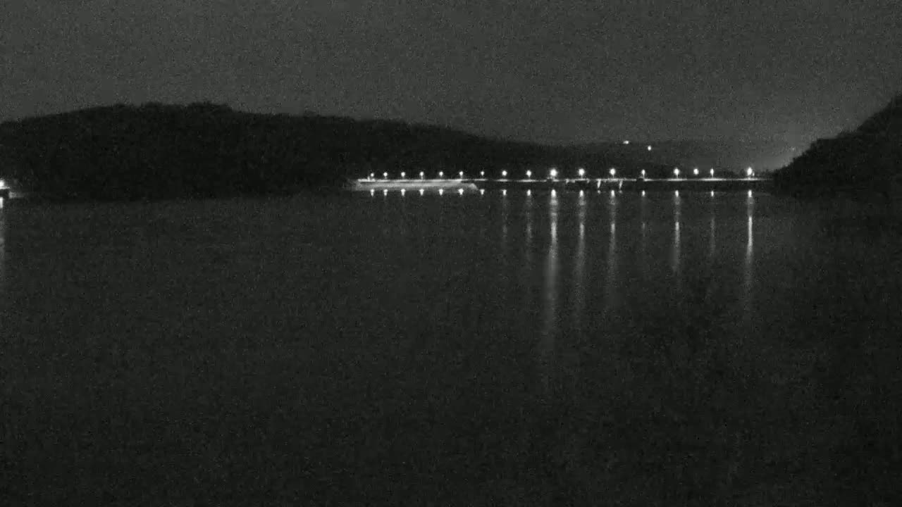 A nighttime view of a calm body of water reflecting the lights of a structure on the far shore, with dark hills in the background.