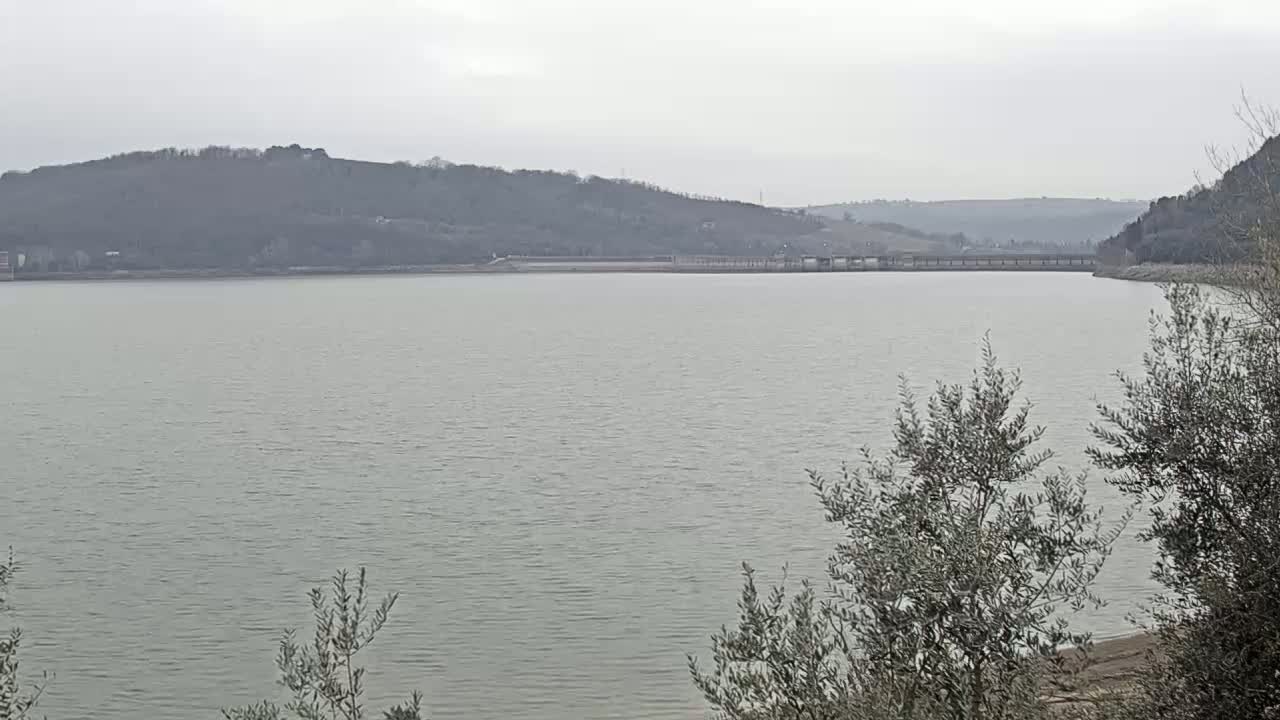 A calm, gray lake stretches out before a line of bare, low hills under a cloudy sky.