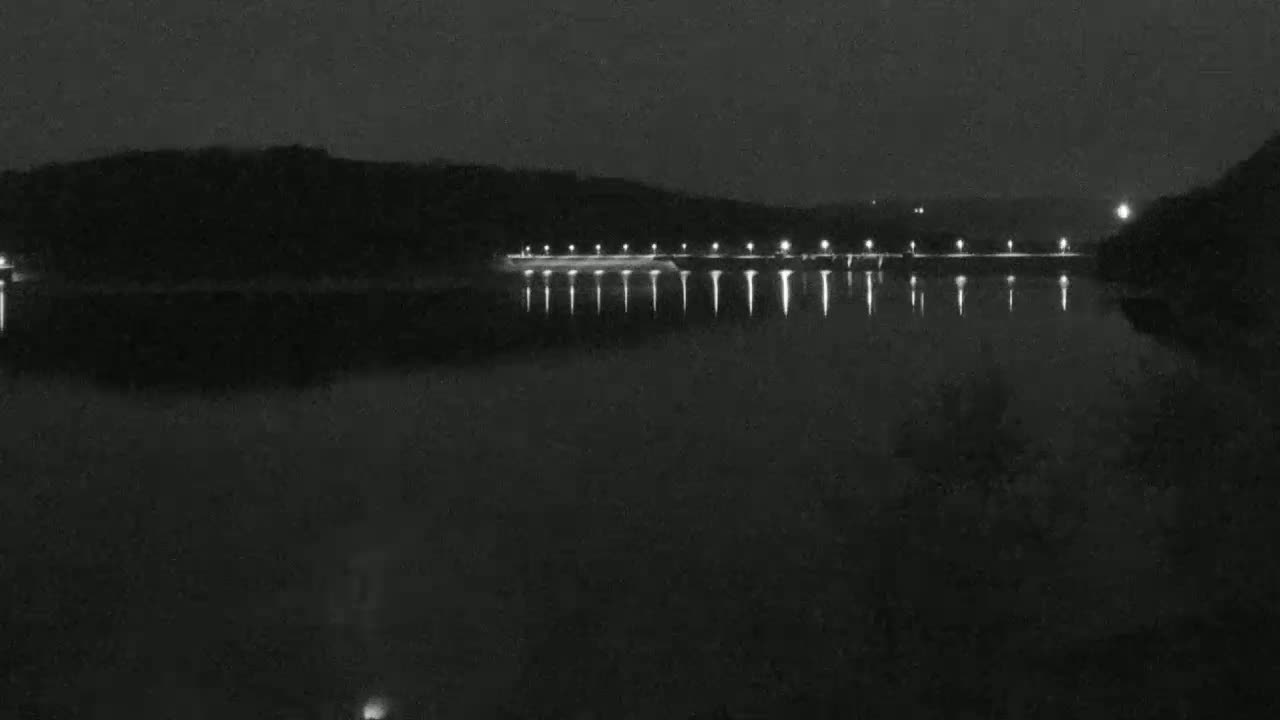 A dark, nighttime scene shows a body of water reflecting the lights of a structure on the far shore, under a clear, moonless sky.