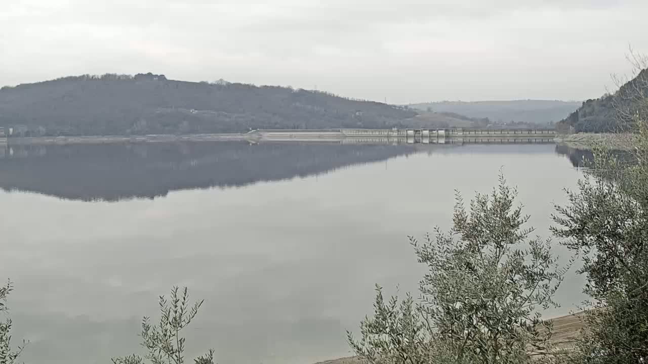 A calm lake reflects a treeline and a long dam under an overcast sky.