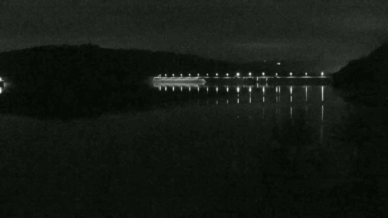 A dark, still body of water reflects the lights of a dam at night.