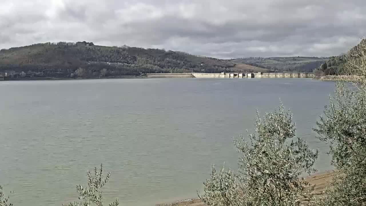 A calm body of water stretches out before a dam, backed by a treeline and rolling hills under a cloudy sky.