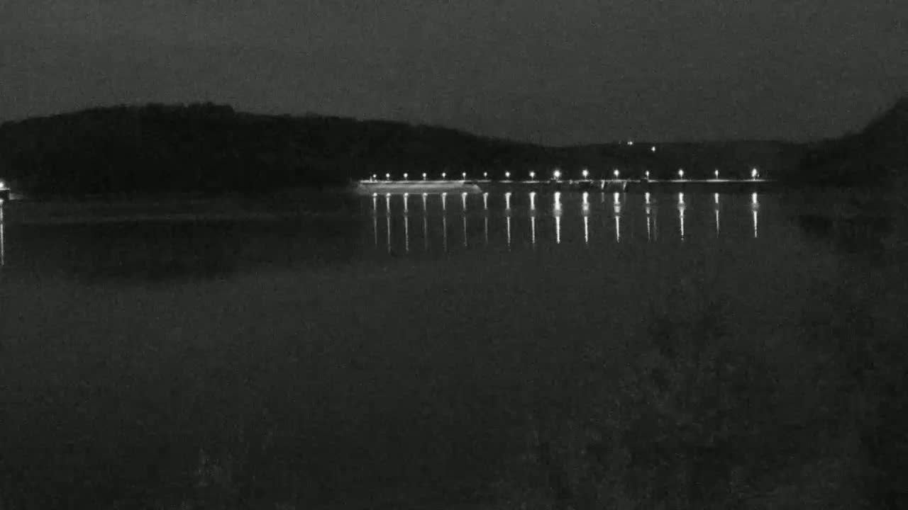 A dark, nighttime scene shows a body of water reflecting the lights of a long structure on the far shore, backed by a dark, wooded hill.