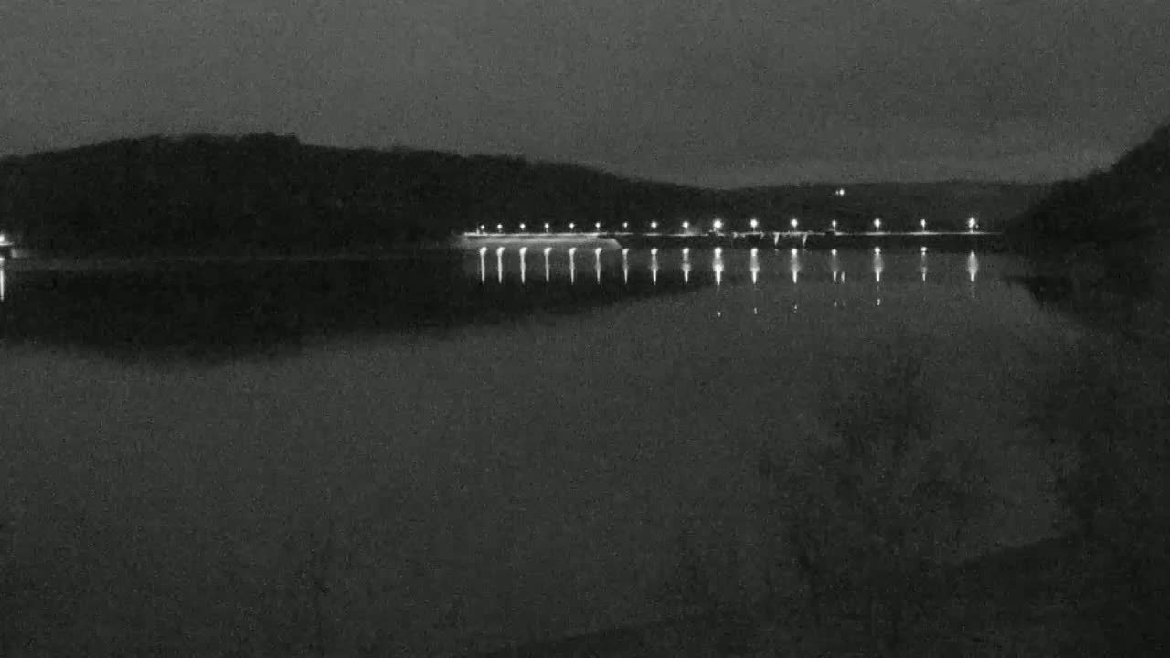 At night, a calm body of water reflects the lights of a dam against a dark, hilly background.