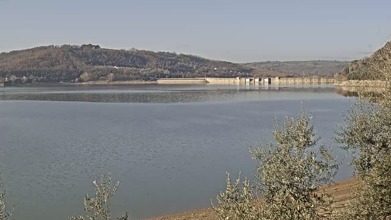 A calm lake stretches out before a dam nestled against a backdrop of low, barren hills under a clear sky.