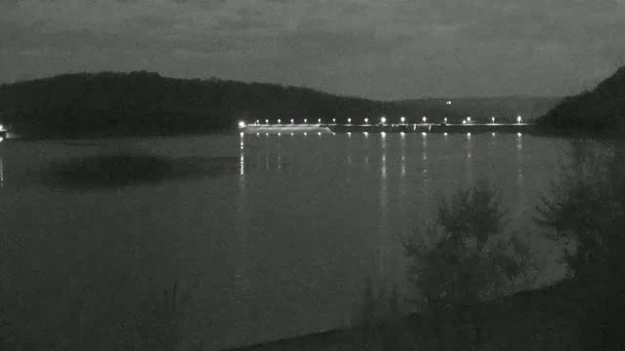 A nighttime view of a calm body of water reflecting the lights of a dam set against a dark, hilly landscape.