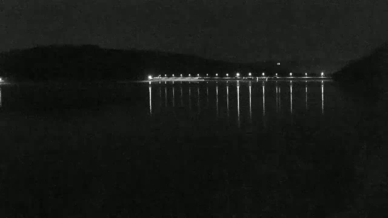 At night, a line of lights reflects on a still body of water in front of a dark hillside.