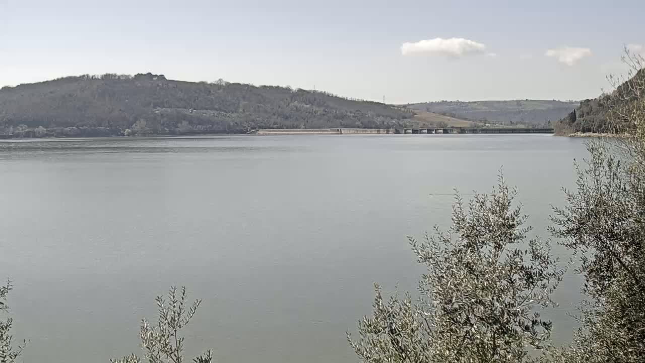 A calm lake stretches out before a hilly landscape under a mostly clear sky.