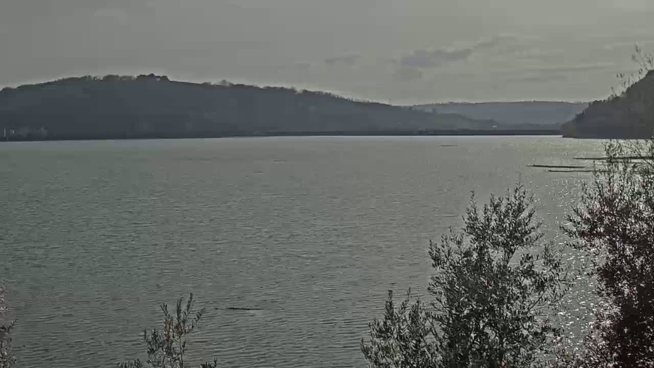 A calm body of water stretches out before a hilly landscape under a mostly cloudy sky.
