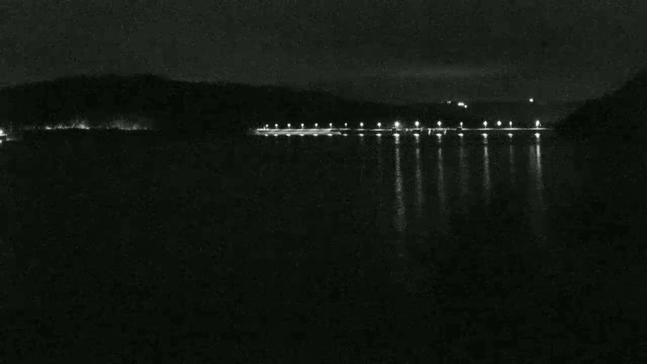 A nighttime view of a calm body of water reflecting the lights of a shoreline, with dark hills in the background under a clear sky.