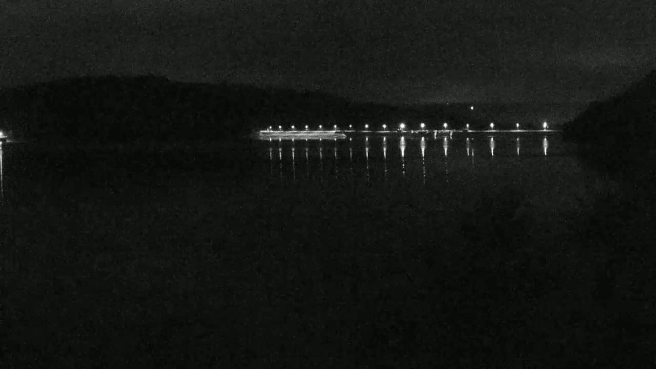 A dark, nighttime scene shows a body of water reflecting the lights of a long, low structure on the far shore under a clear, black sky.