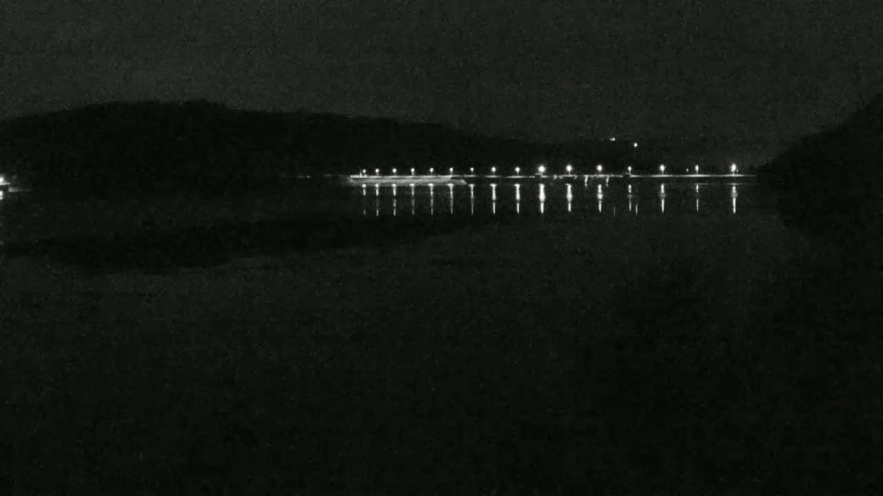 A dark, nighttime scene shows a calm body of water reflecting the lights of a distant shoreline.