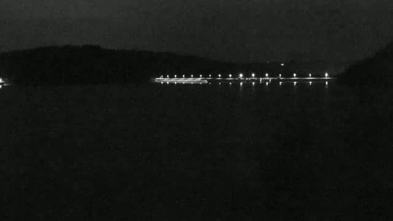 A line of lights illuminates a dark body of water at night, with a dark shoreline in the background.