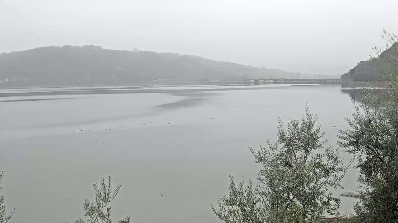 A calm lake stretches out under a foggy sky, with a bridge and distant hills barely visible in the hazy distance.